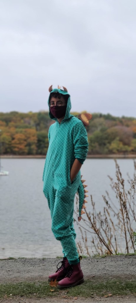Mylo wearing a maroon maks, maroon boots, and a dragon onesie in front of a river on a fall day.